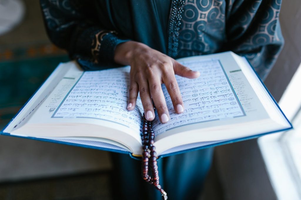 A woman's hand rests on an open Quran with prayer beads, symbolizing devotion.